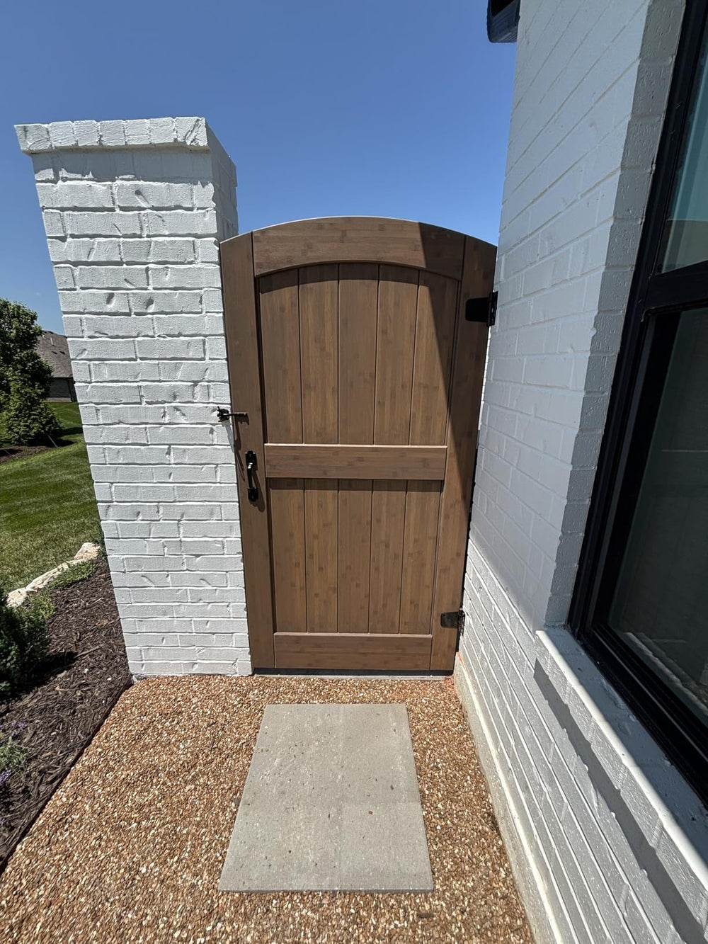 Brown-stained arched bamboo gate beside a white-painted brick pillar